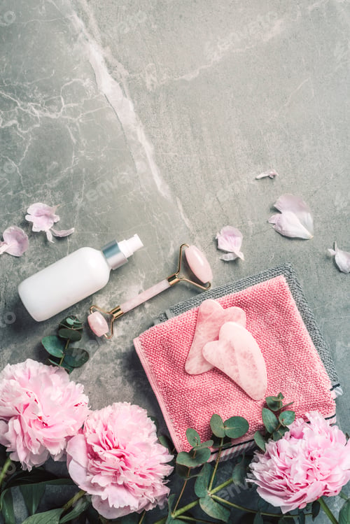 Preview: Facial roller, gua sha massager, oil bottle, towel, pink peonies on marble background. Copy space