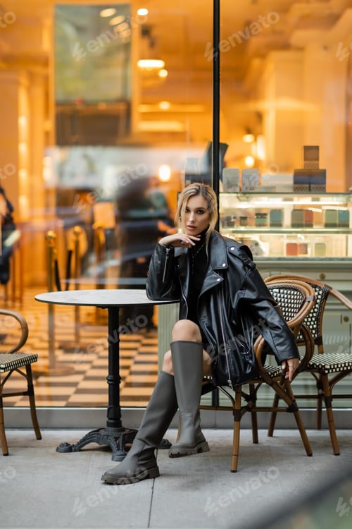 Preview: full length of blonde woman in leather jacket sitting near round bistro table in outdoor cafe in