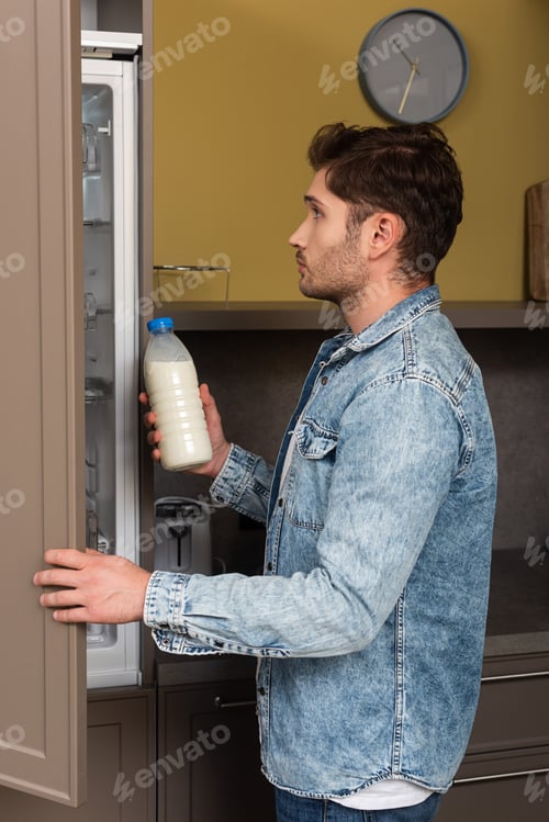 Preview: Side view of handsome man holding bottle of milk near open fridge in kitchen