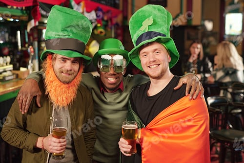 Preview: Three happy young intercultural men in green hats toasting with glasses of beer