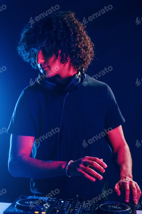 Preview: Man with curly hair using DJ equipment and standing in the dark neon lighted room
