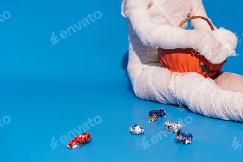 Preview: Child in mummy costume eats candies from basket in the form of pumpkin against blue background