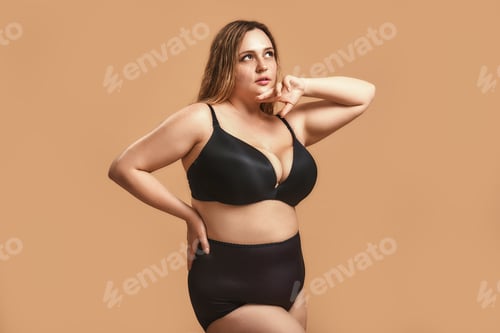 Preview: Chubby pretty woman in sexy black lingerie looking away while standing against brown background in