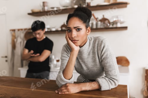 Preview: Upset african american woman leaning on hand sadly looking in camera with asian man using cellphone
