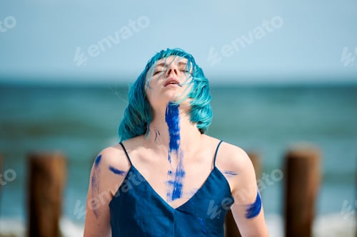 Preview: Artistic blue-haired woman performance artist smeared with blue gouache paints dancing on beach