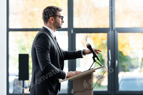 Preview: side view of businessman in suit standing at podium tribune and looking away during conference