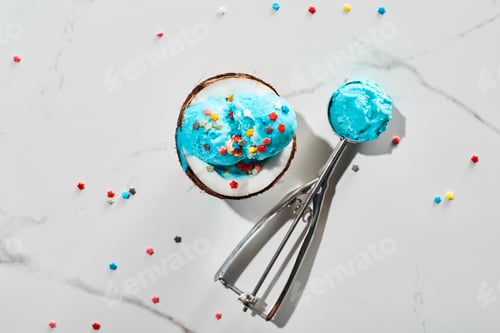 Preview: top view of fresh blue ice cream with sprinkles on coconut half and in ice cream spoon on marble