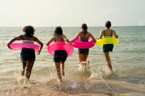 Preview: Back of teen girls with swimming rings run into the sea with warm light of sunset during holiday