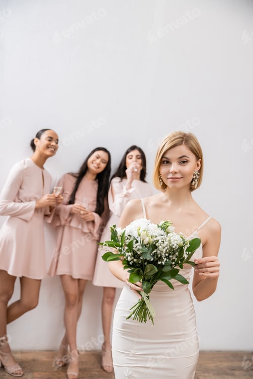Preview: beautiful bride in wedding dress holding bridal bouquet, standing near blurred interracial