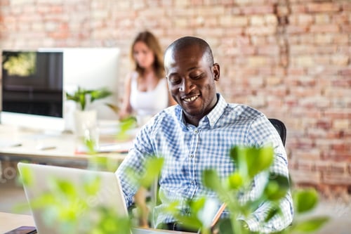 Preview: Smiling businessman using laptop at desk in office with colleague in background