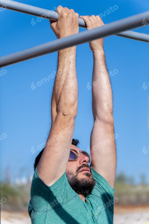 Preview: man with beard and sunglasses training his back by doing pull-ups on a bar in an outdoor gym