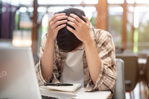 Preview: Man in plaid shirt holding head thinking or stressing in front of laptop and notebook at cafe table.