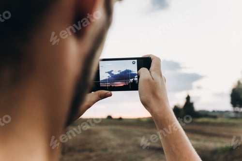 Preview: man holding phone and taking photo of amazing sunset landscape view in summer field