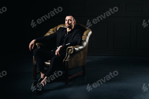 Preview: portrait of an elegant young European man in a suit sitting on a chair in a dark interior