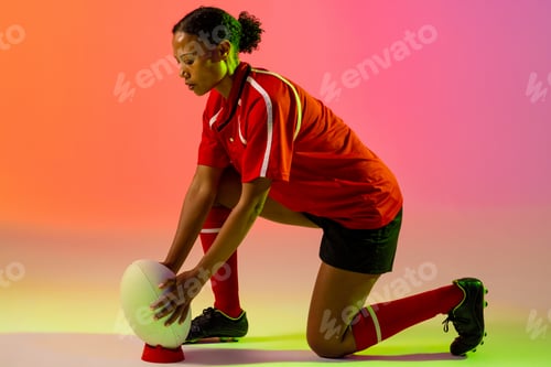 Preview: African american female rugby player crouching with rugby ball over neon pink lighting