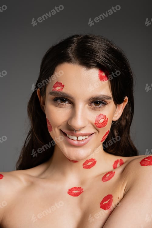 Preview: young and happy woman with red kiss prints on face and bare shoulders looking at camera isolated on