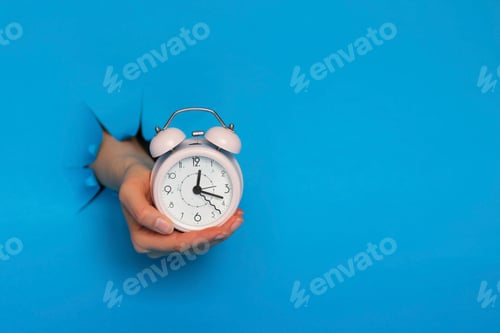 Preview: female hand holds pink alarm clock through a paper hole in blue background.