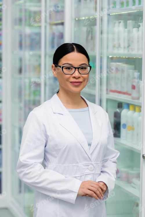 Preview: cheerful asian pharmacist in glasses standing with clenched hands in drugstore