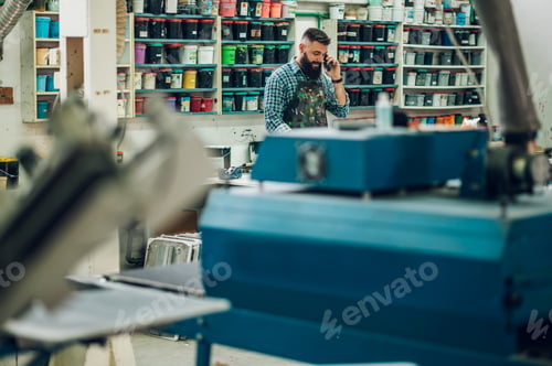 Preview: Male worker holding a screen printing film in a workshop and using a smartphone