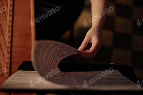 Preview: Female Hand Turning Page with Music Notes while Playing Harp in Low Key Light Indoors
