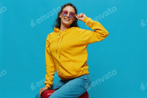Preview: Smile woman sitting on a suitcase in a yellow hoodie, blue jeans and glasses on a blue background