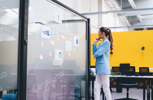 Preview: Young pensive woman standing by glass wall with sticky notes in office