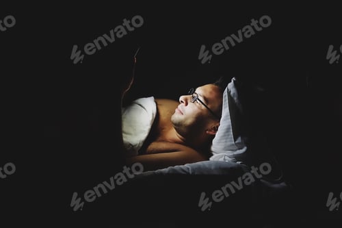 Preview: Man wearing glasses using his mobile device in bed before sleep with light from the phone