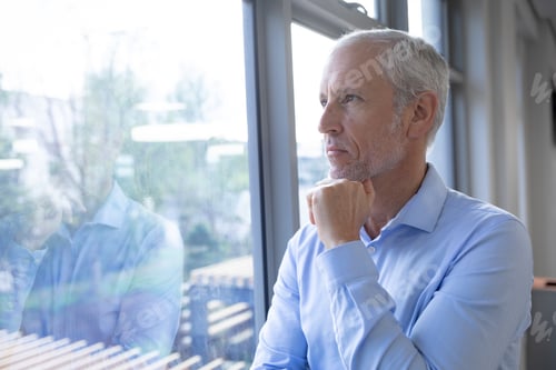 Preview: Senior businessman with hand on his chin looking through window at modern office