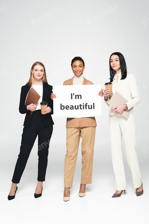 Preview: happy multicultural businesswomen holding folders and paper cups near placard with i`m beautiful