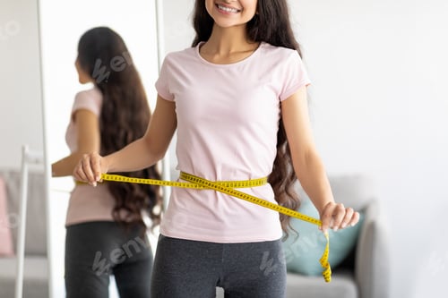 Preview: Unrecognizable Indian lady measuring her waist with tape in front of mirror at home, closeup