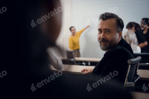 Preview: Mature guy turns to consult with attendees sitting at a table in the back of the conference room