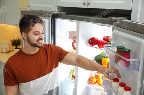 Preview: Young man taking yoghurt out of refrigerator indoors
