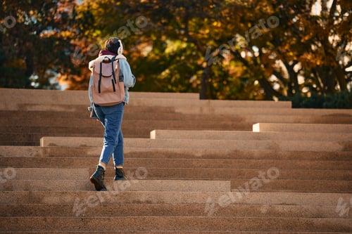 Preview: Rear view of woman with backpack and headphones walking up the stairs outdoors.