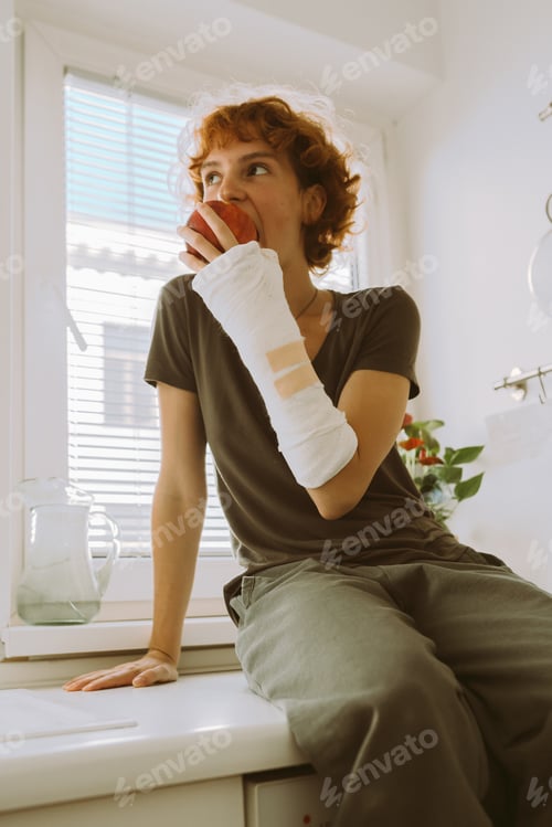 Preview: teenage girl with broken arm in cast in kitchen sitting on countertop eating apple