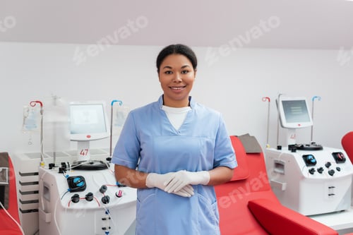 Preview: cheerful multiracial nurse in blue uniform and latex gloves looking at camera near automated