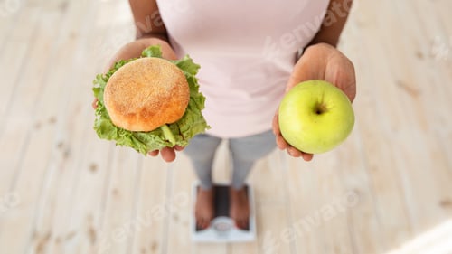 Preview: Above view of black woman holding apple and hamburger, standing on scales, making choice of her diet