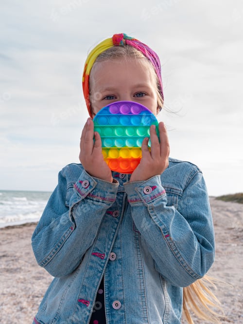 Preview: Blond child girl rainbow toy walking having fun on beach Bright colorful trendy antistress push toy