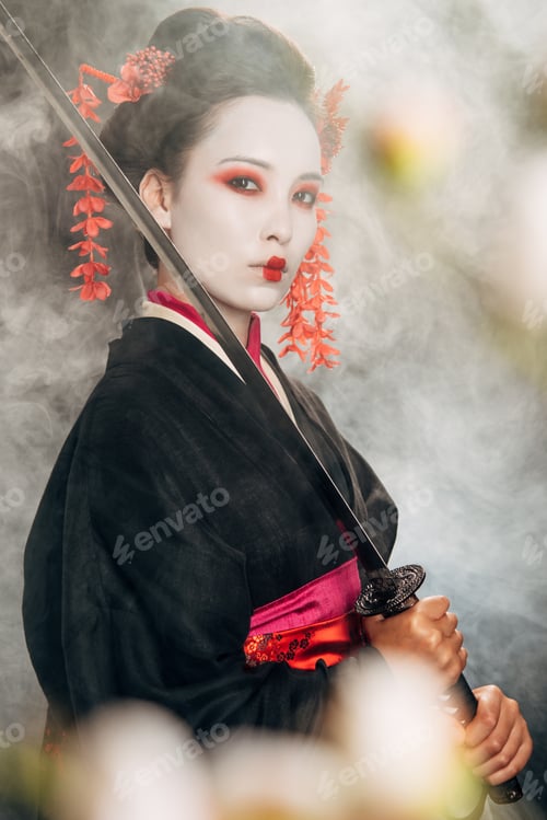 Preview: selective focus of serious geisha in black kimono holding katana in smoke and sakura branches