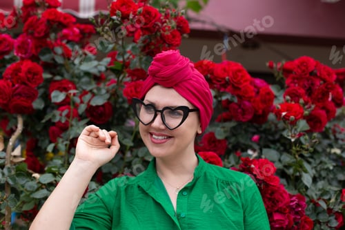 Preview: Candid portrait of beautiful vibrant woman in bright clothes and funny glasses by red roses bush