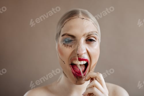 Preview: Adult woman screams violently. Portrait of girl tearing up mask with terrible makeup