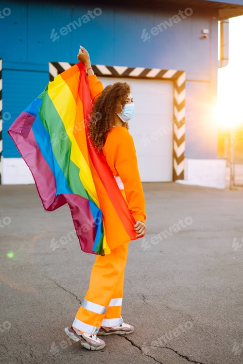 Preview: African lesbian woman with medical face mask posing with lgbt pride flag. Woman during quarantine.
