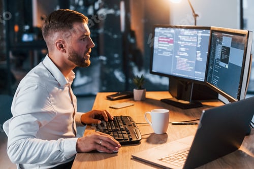 Preview: Night time. Young businessman in formal clothes is in office with multiple screens