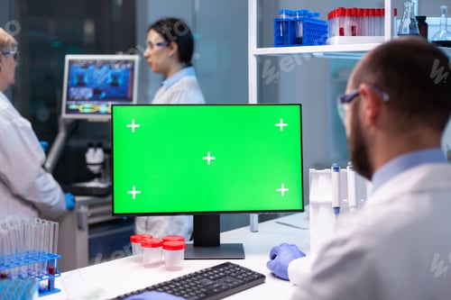 Preview: Biochemist sitting at workplace in modern laboratory using green mock-up screen