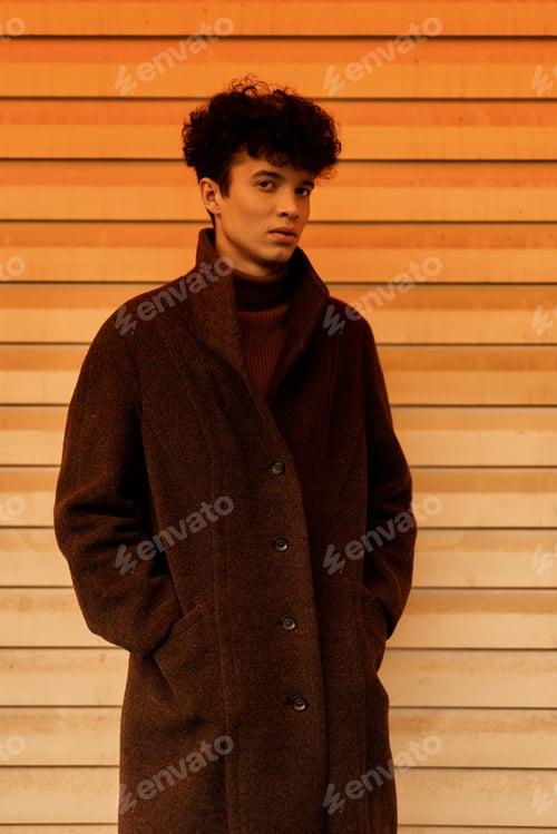 Preview: Fashionable young man in a brown coat posing in front of a wooden wall with orange and white stripes