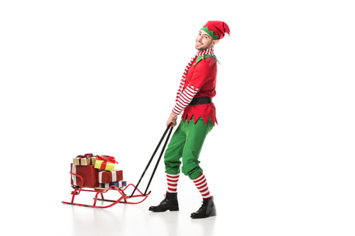 Preview: Man in christmas elf costume crrying pile of presents on sleigh isolated on white