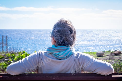 Preview: Rear view of mature woman relaxing in front to the sea, sitting on a bench in a windy day