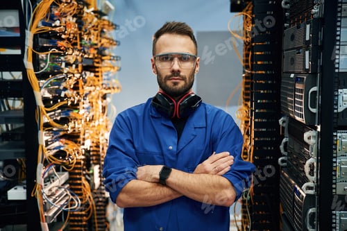 Preview: Standing with arms crossed. Young man is working with internet equipment and wires in server room