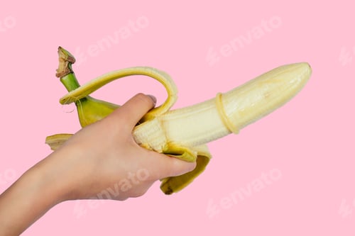 Preview: close up of woman's hands putting banana on condom cock on a pink background