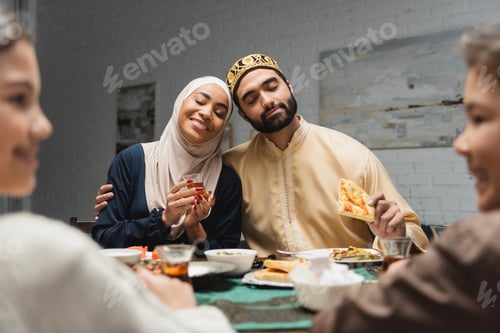 Preview: Arabian man hugging wife in hijab near blurred kids during iftar at home