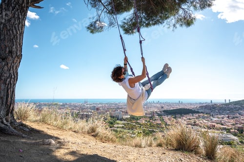Preview: Caucasian woman riding on a swing on the edge of a hill with the background of a townscape